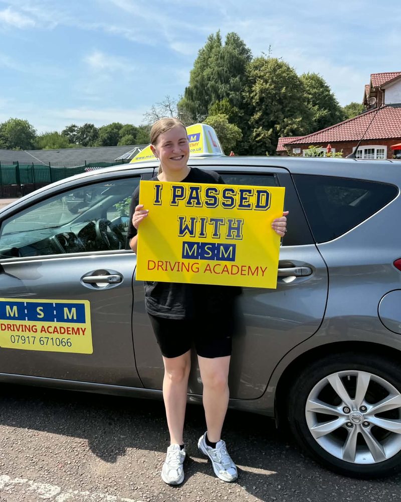 A smiling learner holding a yellow 'I passed with MSM Driving Academy' board in front of a grey Nissan Note tuition car on a sunny day