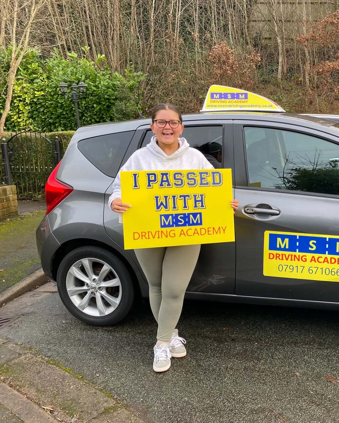 A smiling young woman in a white hoodie holding a yellow 'I passed with MSM Driving Academy' board next to a grey Nissan Note driving-school car