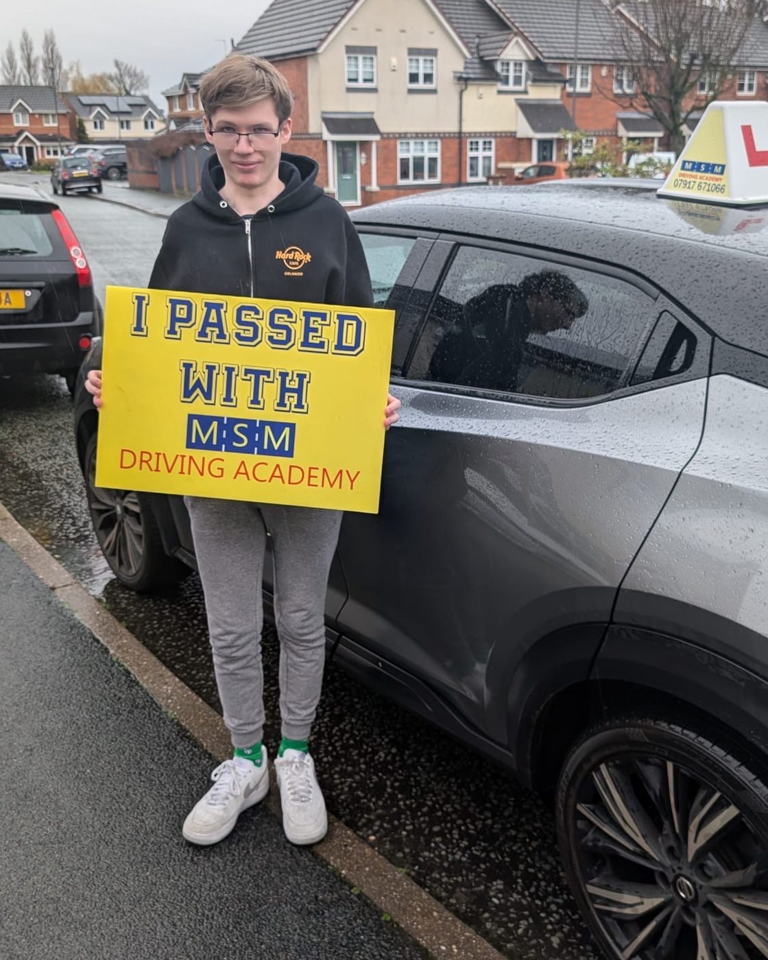 A young man in glasses holding a yellow 'I passed with MSM Driving Academy' board beside a grey tuition car with an L-plate roof sign on a wet residential street