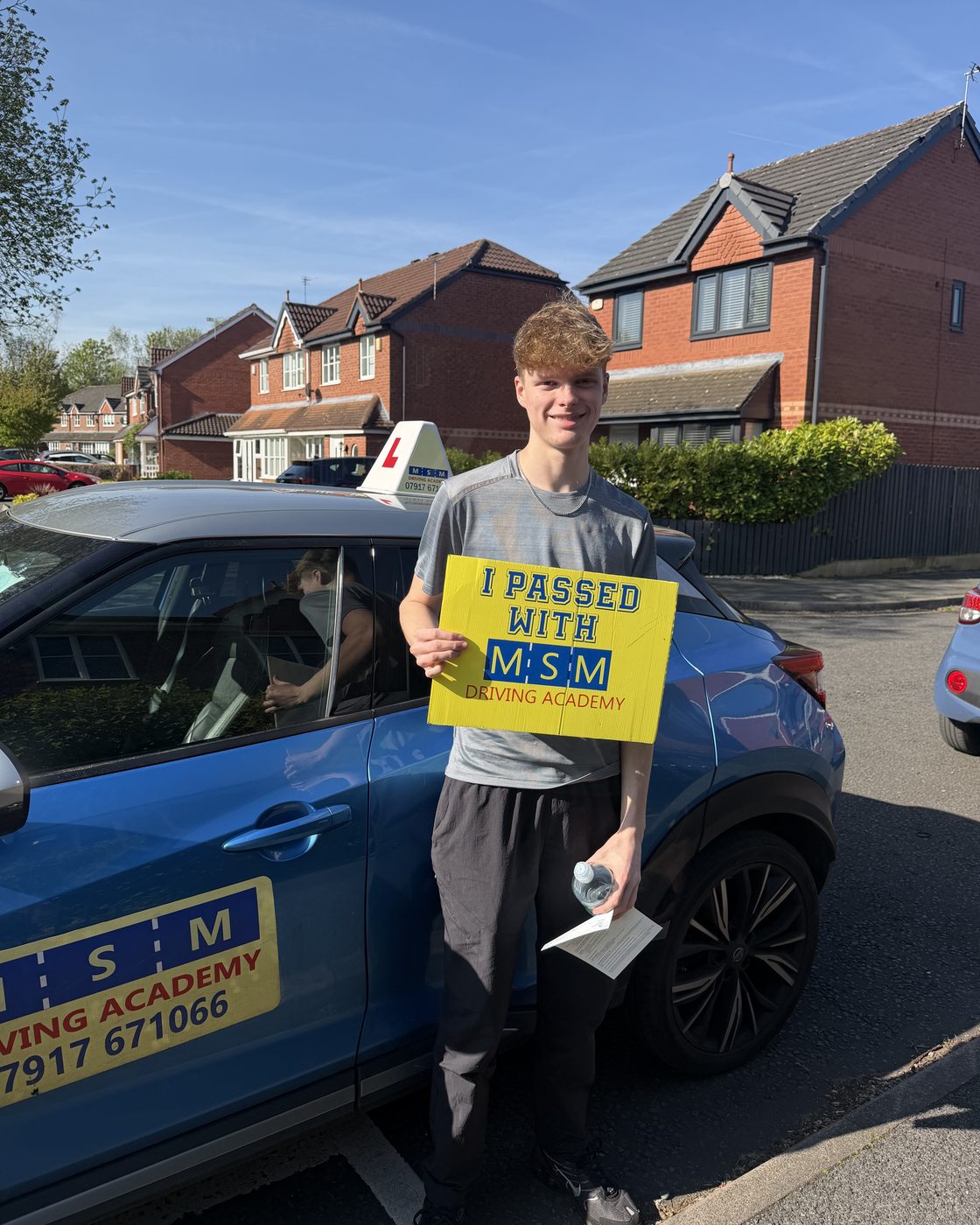 A young man holding a yellow 'I passed with MSM Driving Academy' board next to a blue MSM Nissan Juke on a sunny residential street