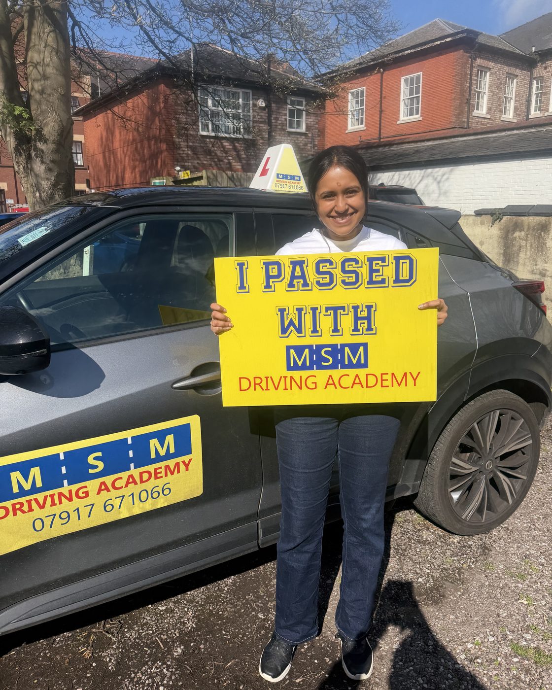 A young woman smiling as she holds a large yellow 'I passed with MSM Driving Academy' board beside a grey tuition car with an L-plate roof sign