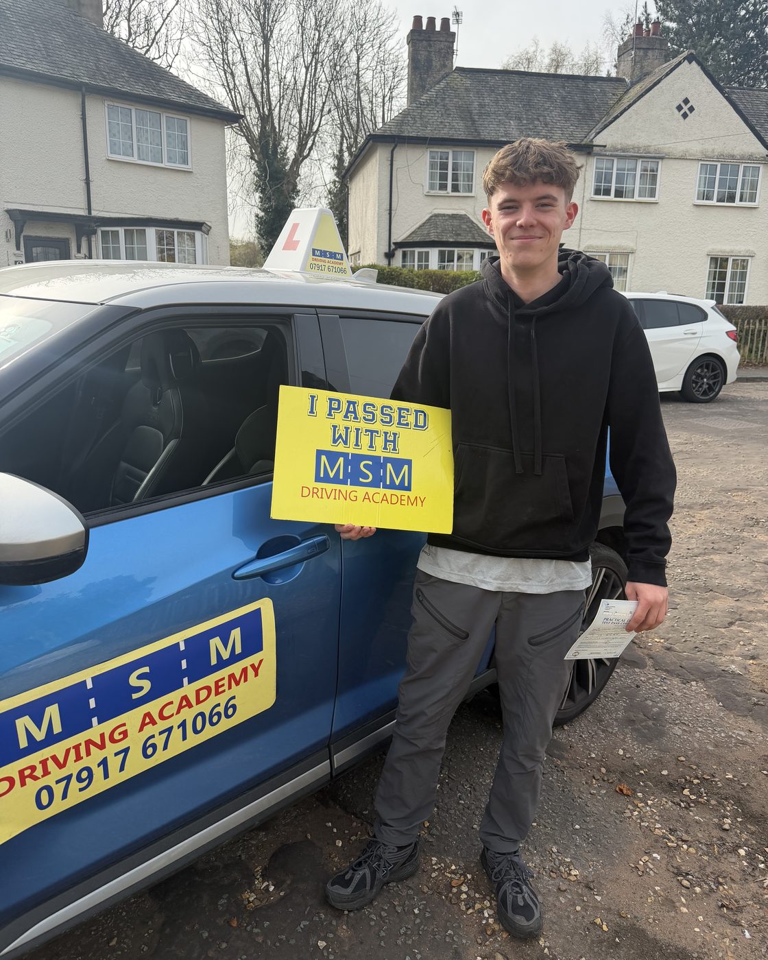 A teenage boy holding a yellow 'I passed with MSM Driving Academy' board next to a blue MSM Nissan Juke outside a row of white cottages