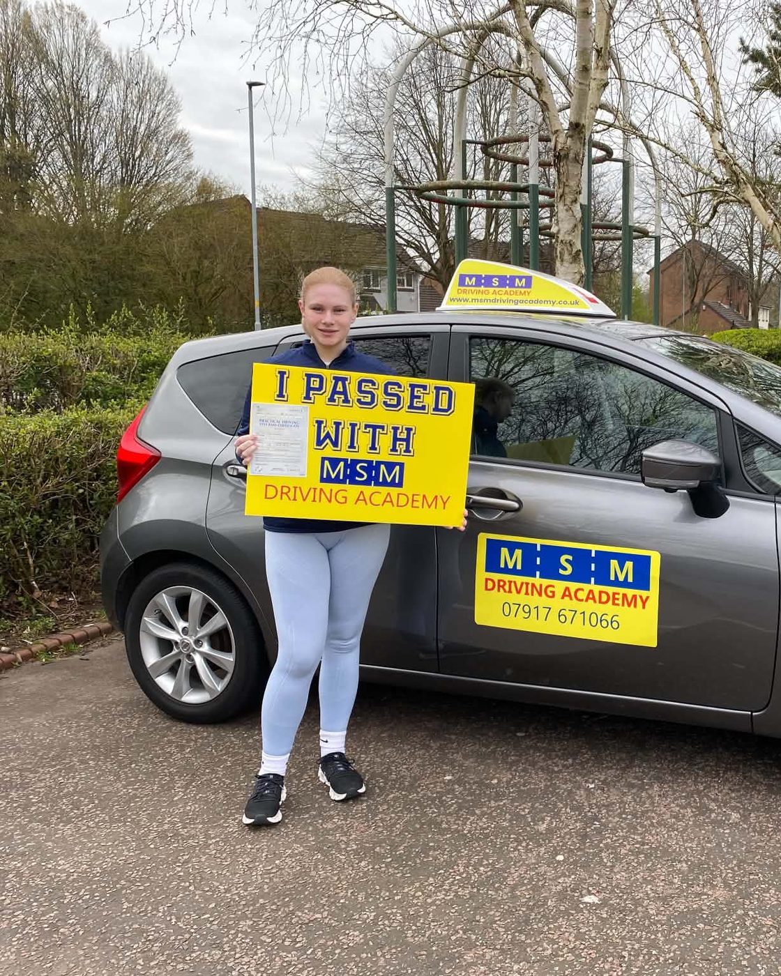 A learner holding a yellow 'I passed with MSM Driving Academy' board beside a grey Nissan Note tuition car with an MSM roof sign