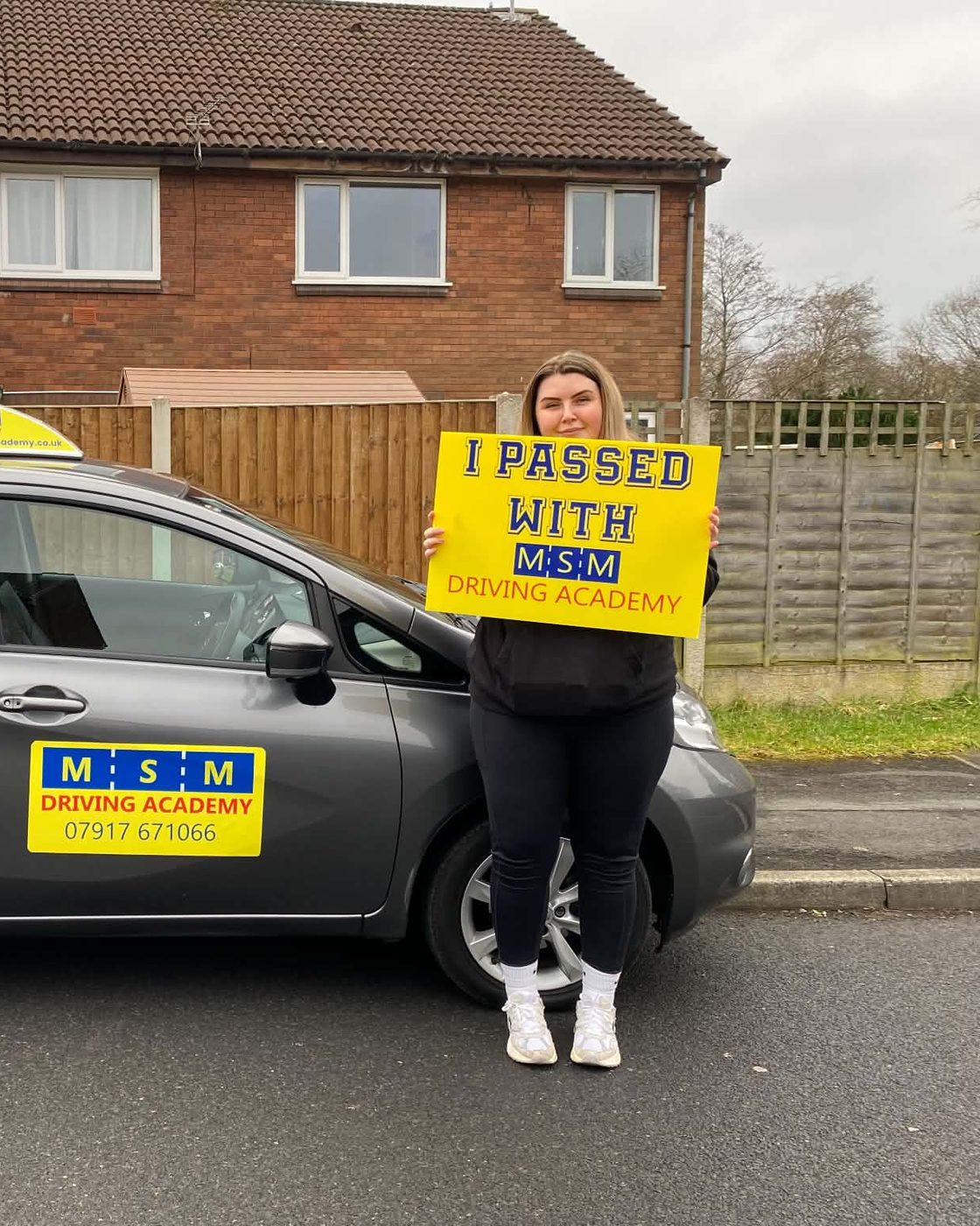 A young woman holding a large yellow 'I passed with MSM Driving Academy' board beside a grey Nissan Note tuition car with an MSM roof sign