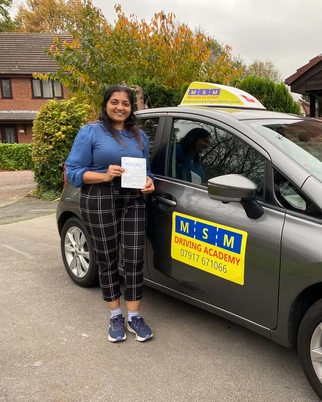 A learner driver smiling and holding up her practical driving test pass certificate beside the grey MSM Driving Academy tuition car