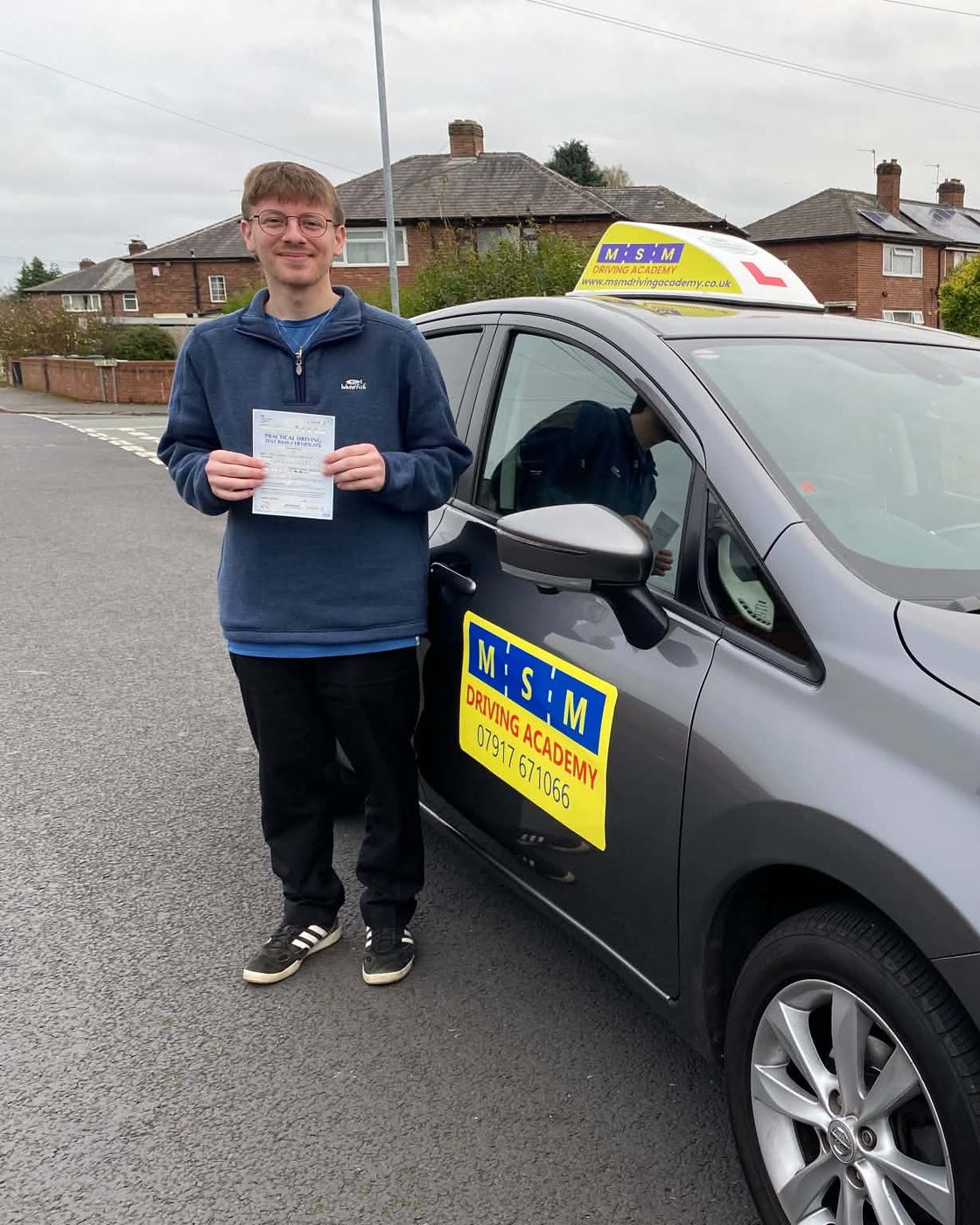 A young man in a blue fleece holding up his practical driving test pass certificate beside a grey Nissan Note MSM tuition car on a residential street