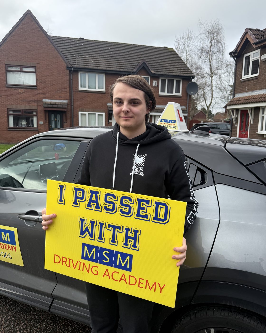 A young man in a black hoodie holding a yellow 'I passed with MSM Driving Academy' board beside a grey tuition car on a residential street