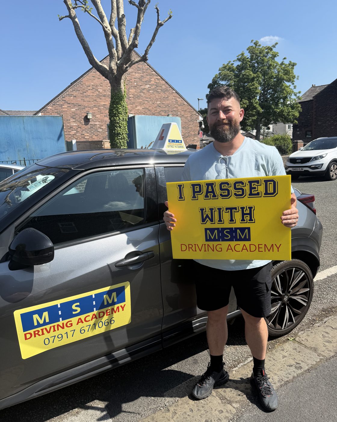 A man in a grey t-shirt holding a yellow 'I passed with MSM Driving Academy' board beside a dark MSM tuition car on a residential street