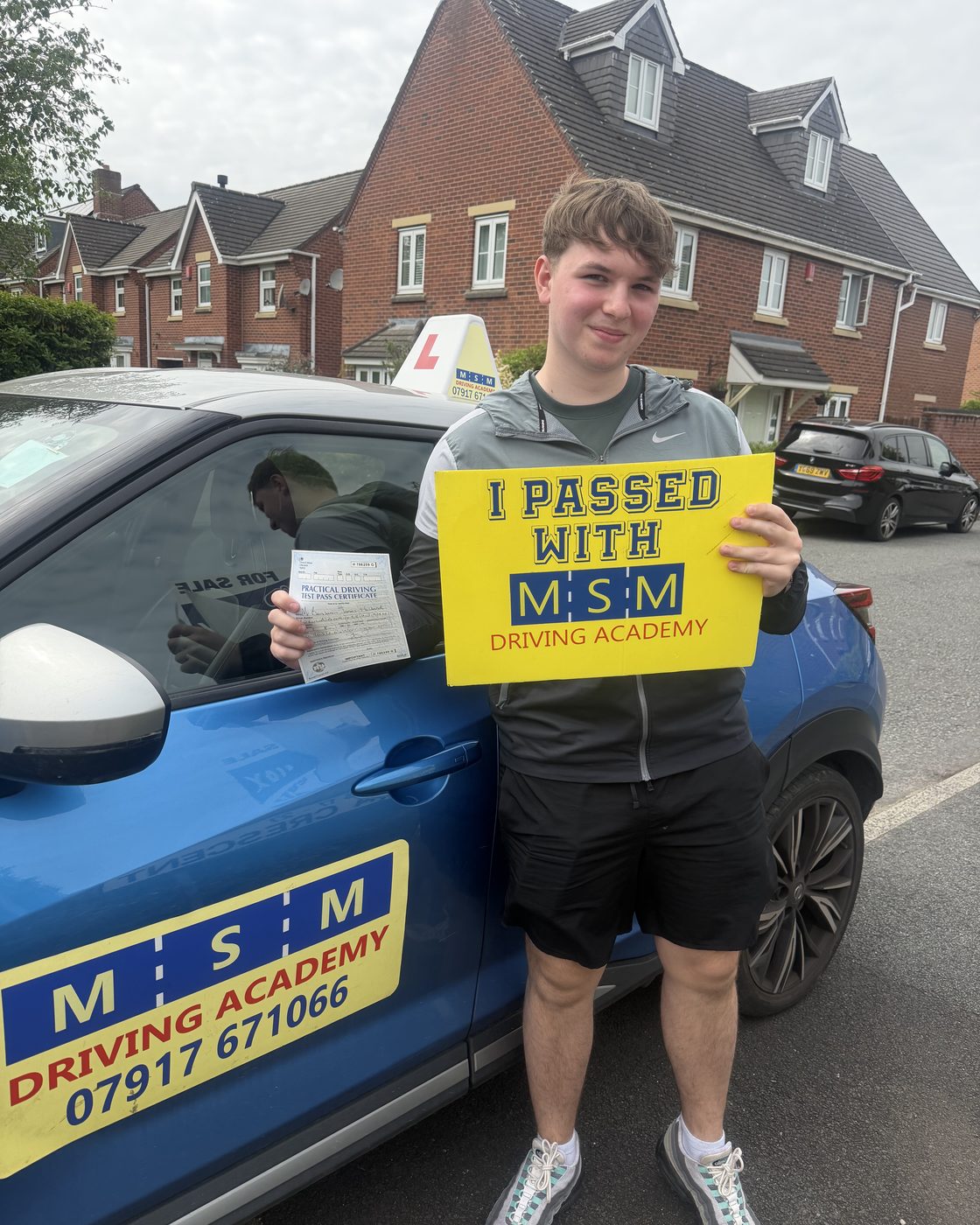 A young man holding up his practical driving test pass certificate and a yellow 'I passed with MSM Driving Academy' board beside the blue MSM Nissan Juke