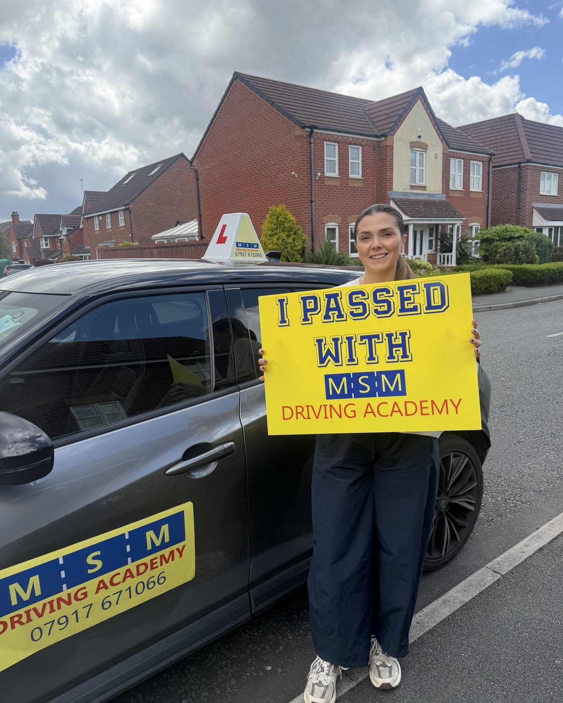 A young woman holding a large yellow 'I passed with MSM Driving Academy' board beside a grey MSM tuition car outside modern houses