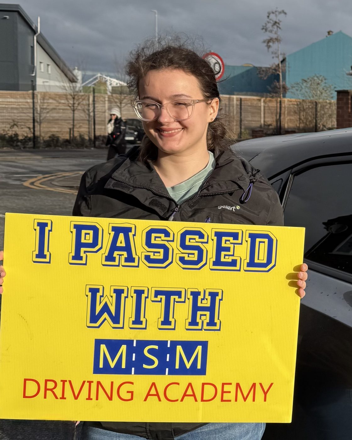 A young woman in glasses smiling behind a large yellow 'I passed with MSM Driving Academy' board beside a dark MSM tuition car