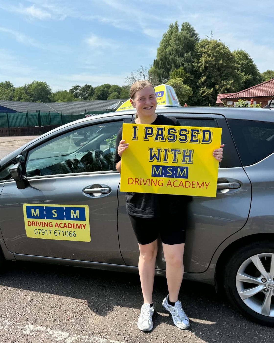 A young woman holding a yellow 'I passed with MSM Driving Academy' board beside the grey Nissan Note automatic tuition car