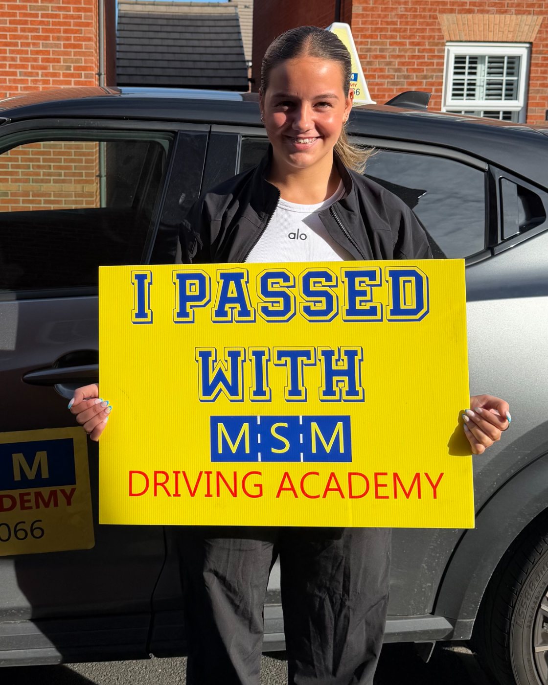 A smiling young woman in a black jacket holding a large yellow 'I passed with MSM Driving Academy' board beside a dark grey MSM tuition car on a sunny residential street