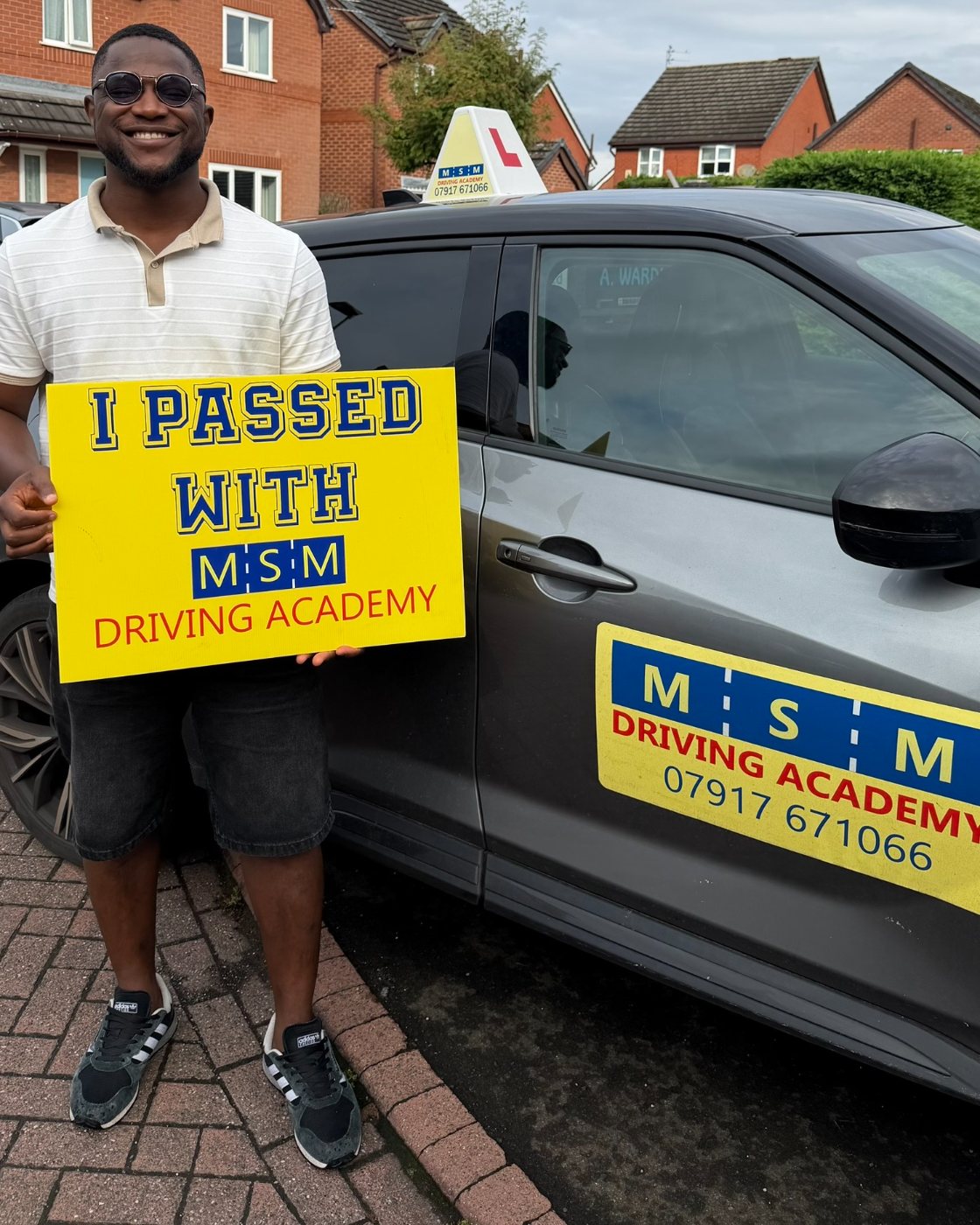 A smiling man in sunglasses and a striped polo shirt holding a large yellow 'I passed with MSM Driving Academy' board beside the grey MSM tuition car